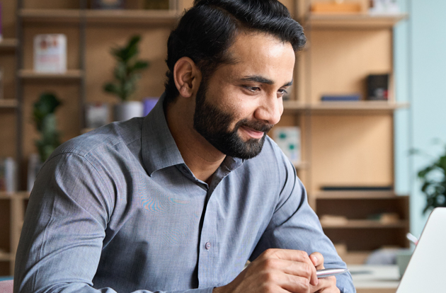 Smiling bearded indian businessman working on laptop at home office. Young indian student using computer remote studying, virtual training on video call meeting, watching online webinar or seminar. People Using Mobile Devices