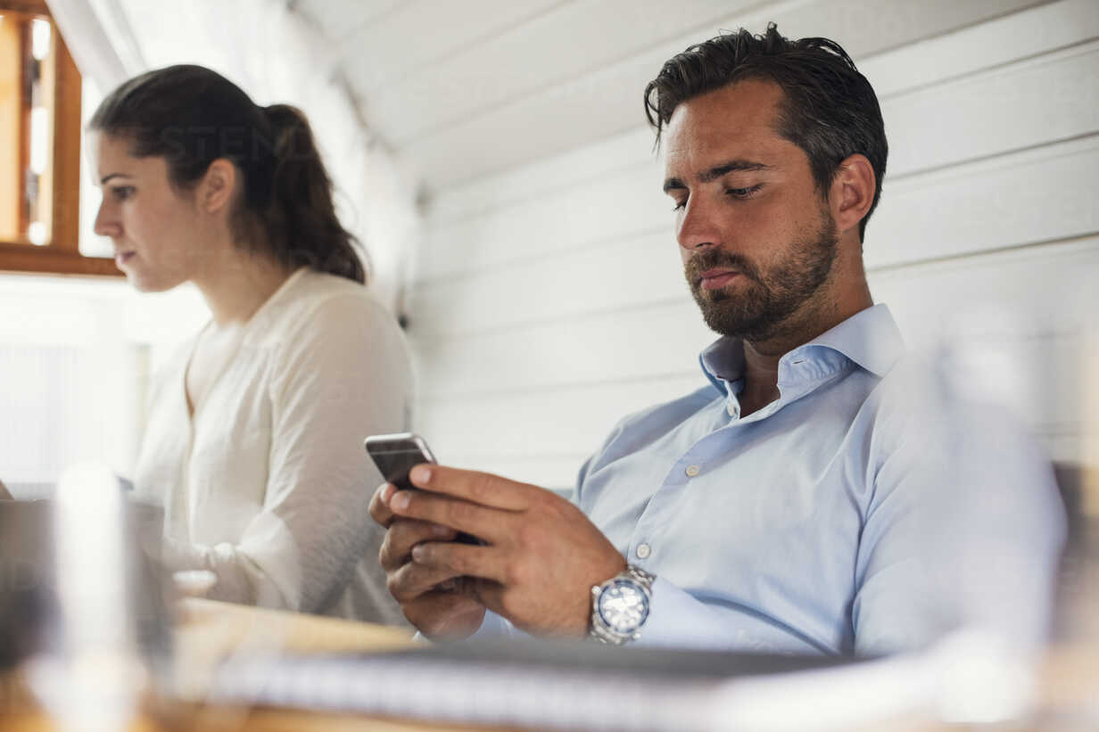 Mid adult businessman using mobile phone by female colleague in office Mid adult businessman using mobile phone by female colleague in office